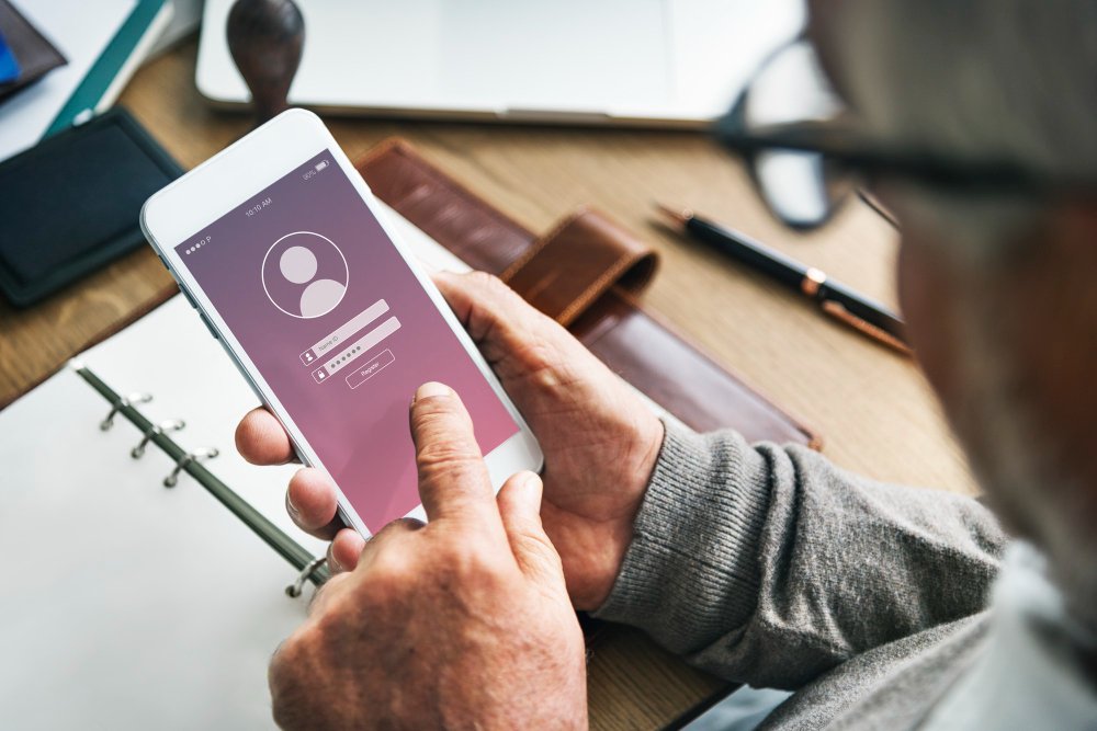Person using smartphone to log into a social media account, featuring a login interface with fields for username and password, surrounded by a workspace with a notebook and pen.